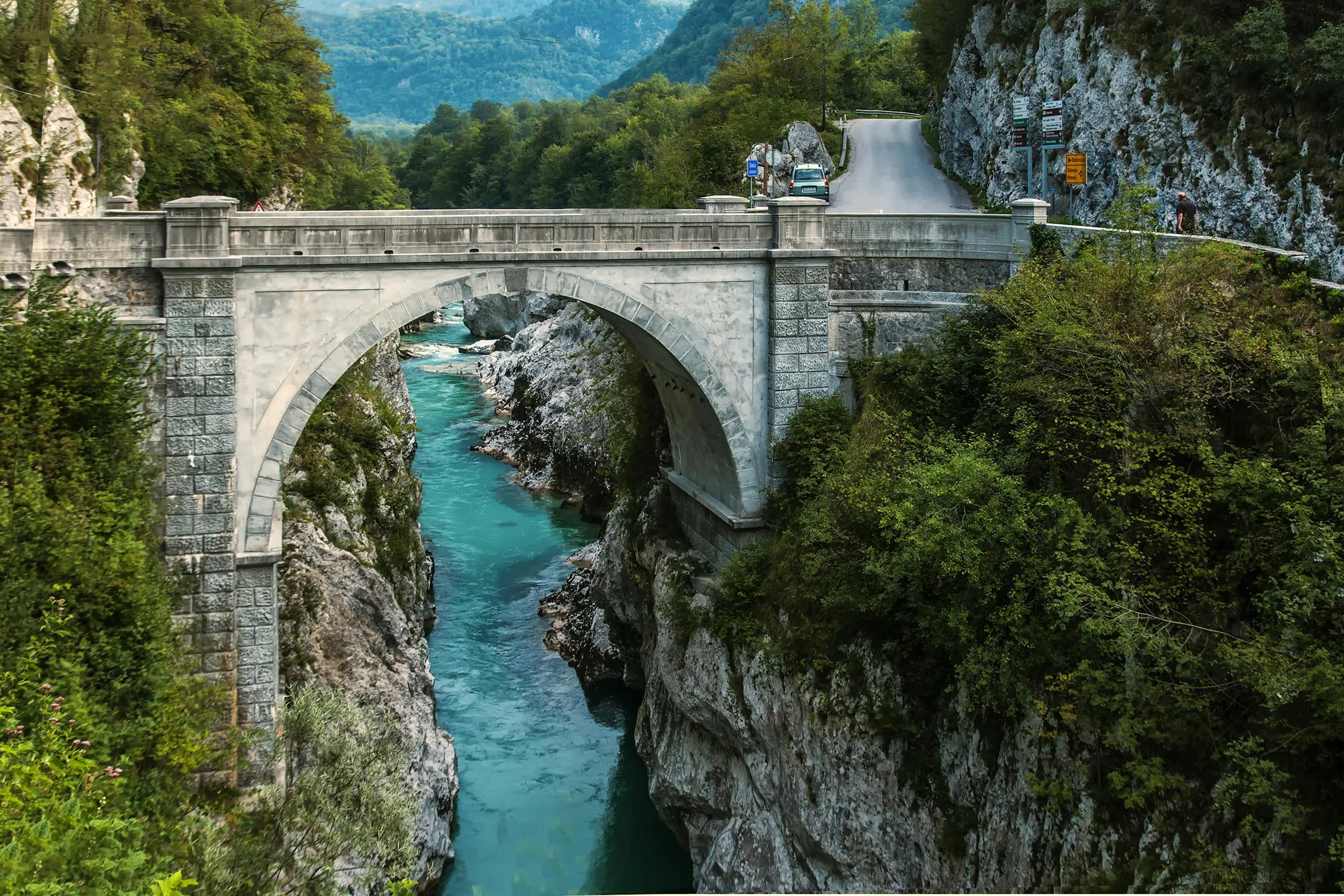 Scenic view of a stone arch bridge over a vibrant turquoise river framed by lush greenery.